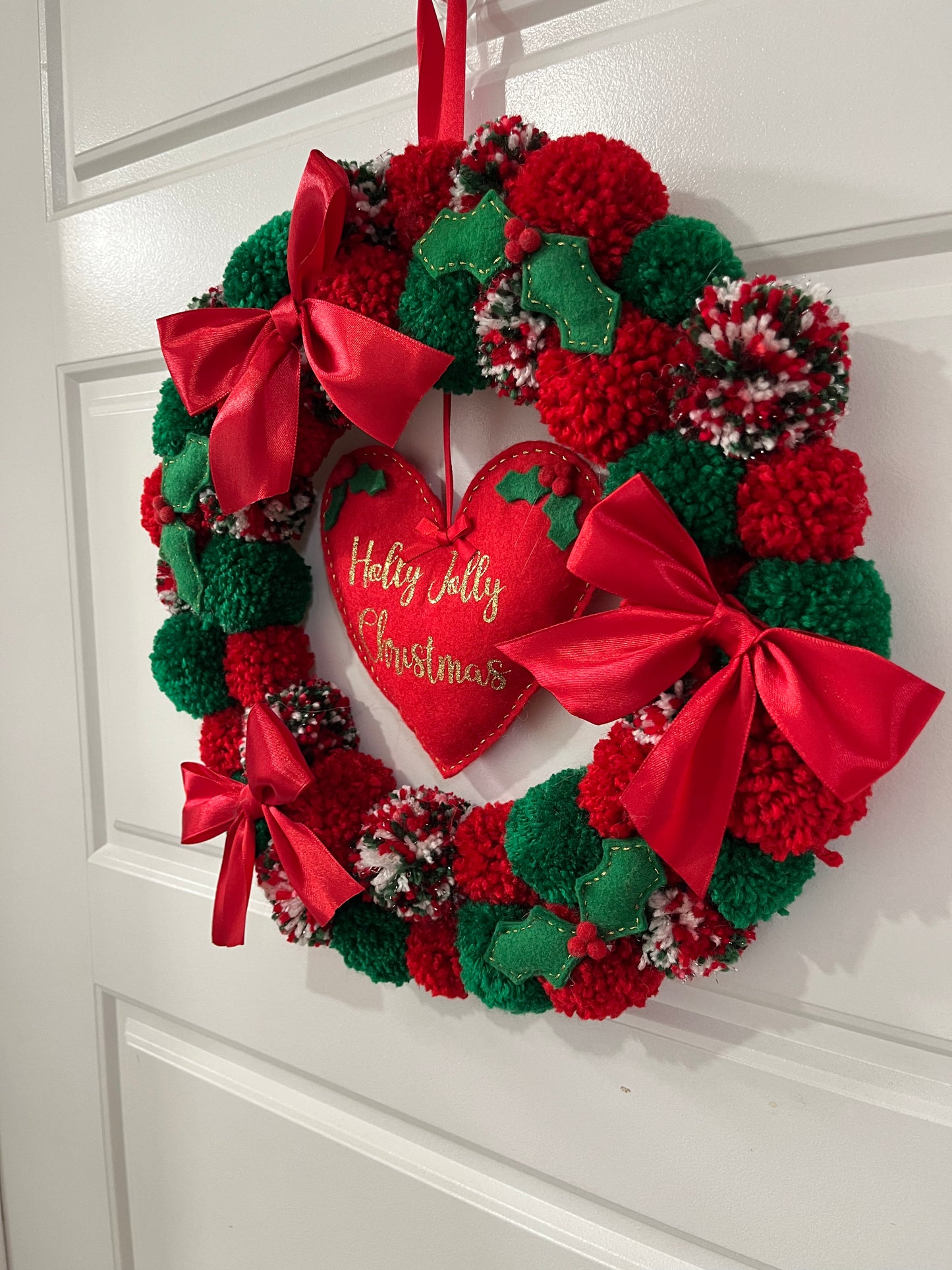 A delightful Christmas wreath decorated with colorful pompoms and a central heart ornament.