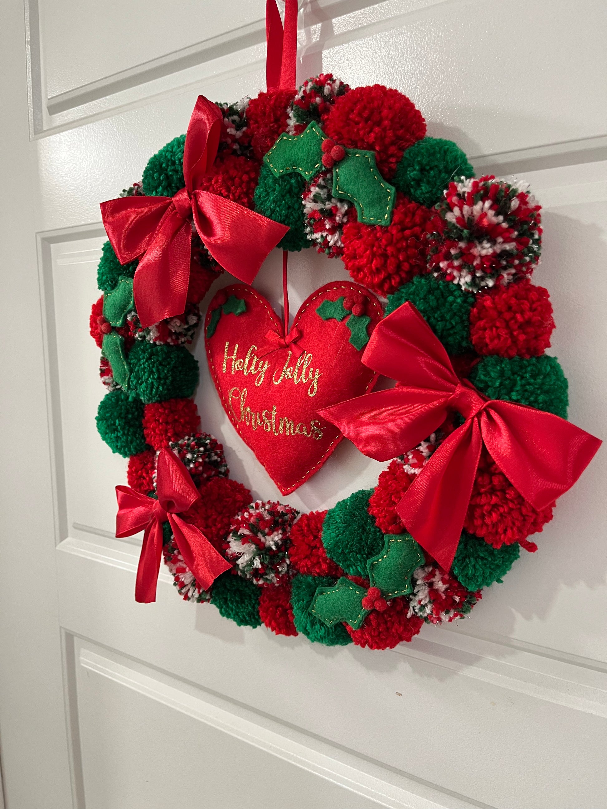 A delightful Christmas wreath decorated with colorful pompoms and a central heart ornament.