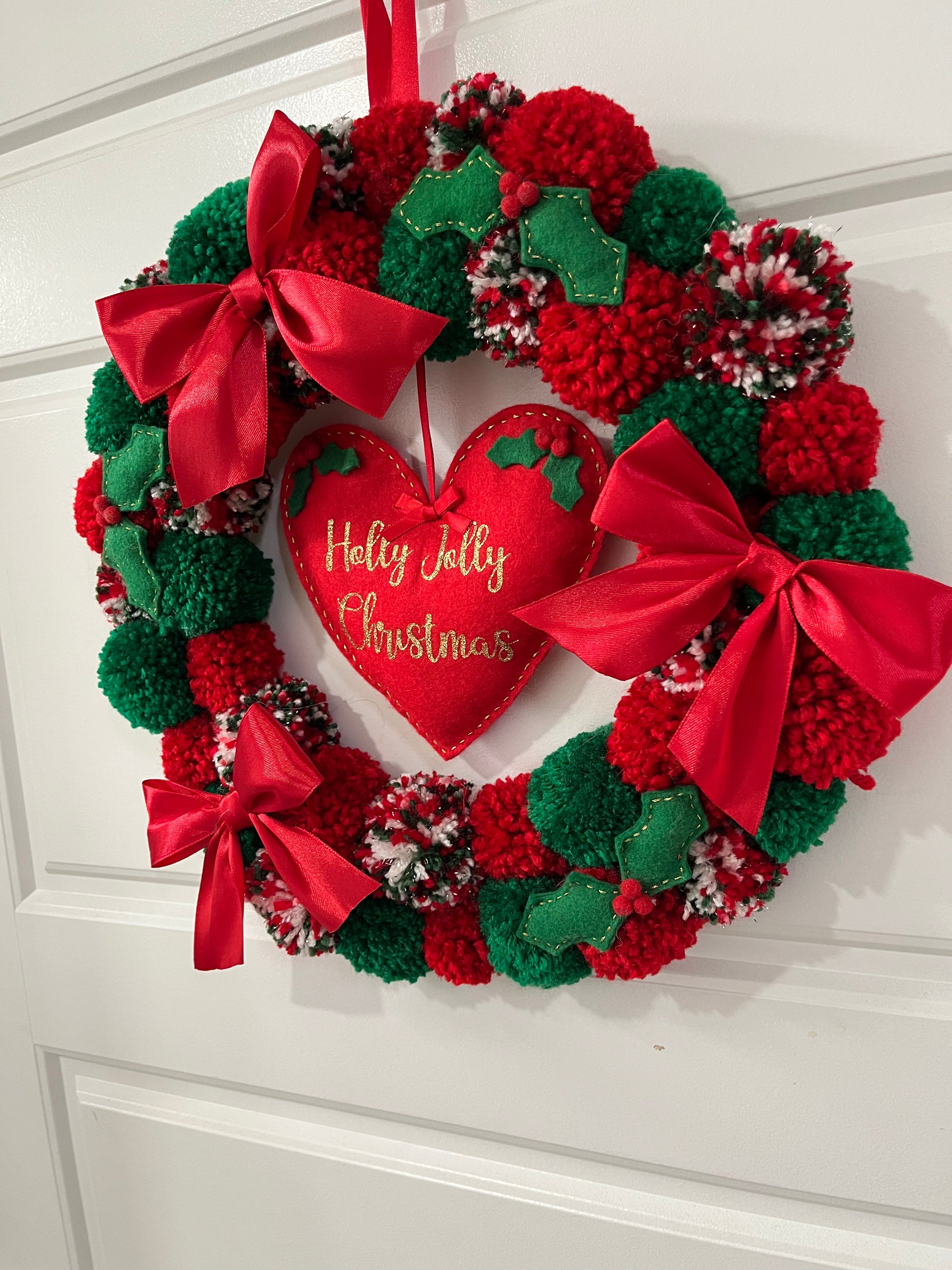 A vibrant Christmas wreath adorned with red and green pompoms, ribbons, and a heart.