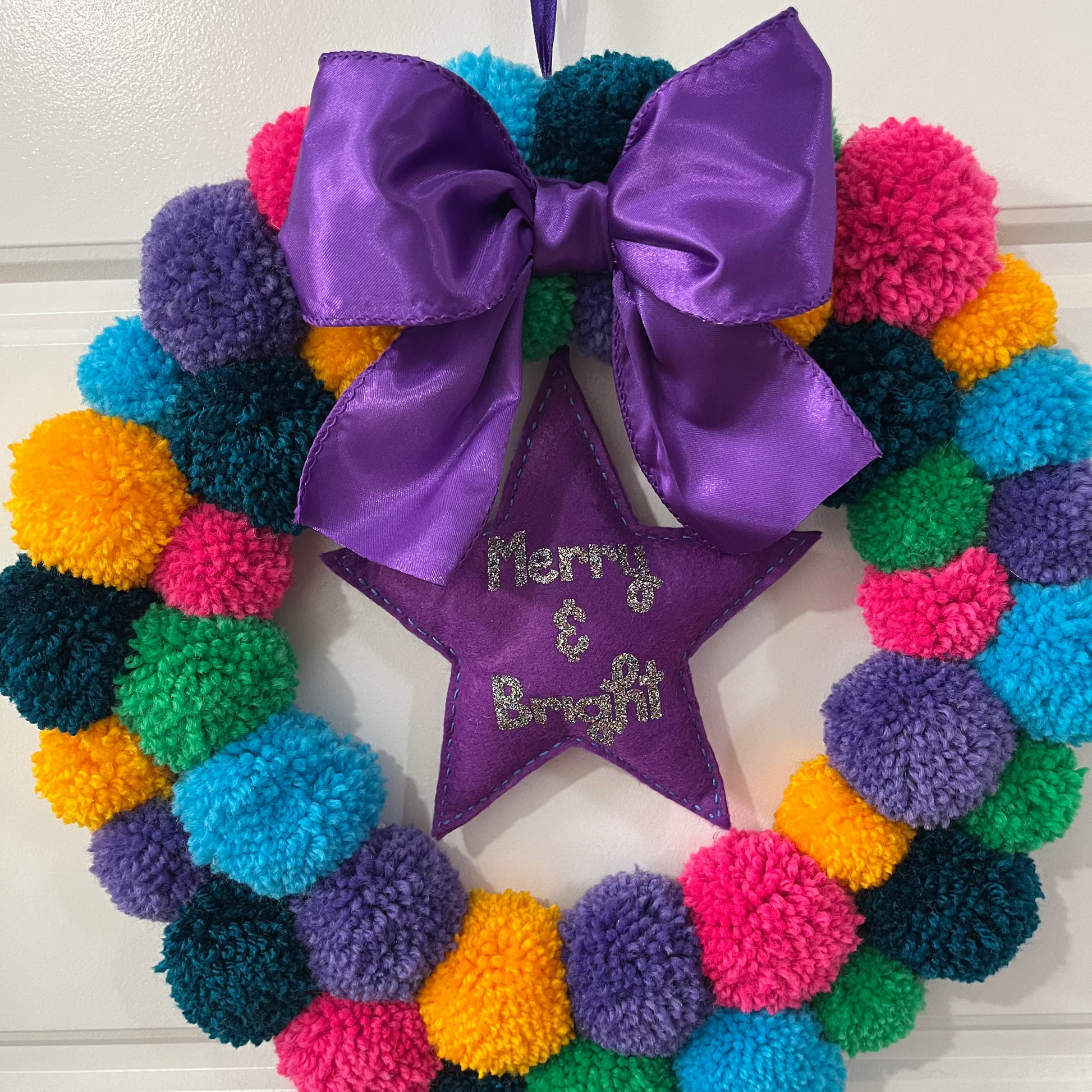 A colorful pom-pom wreath with a purple bow and a star reading 'Merry & Bright'.