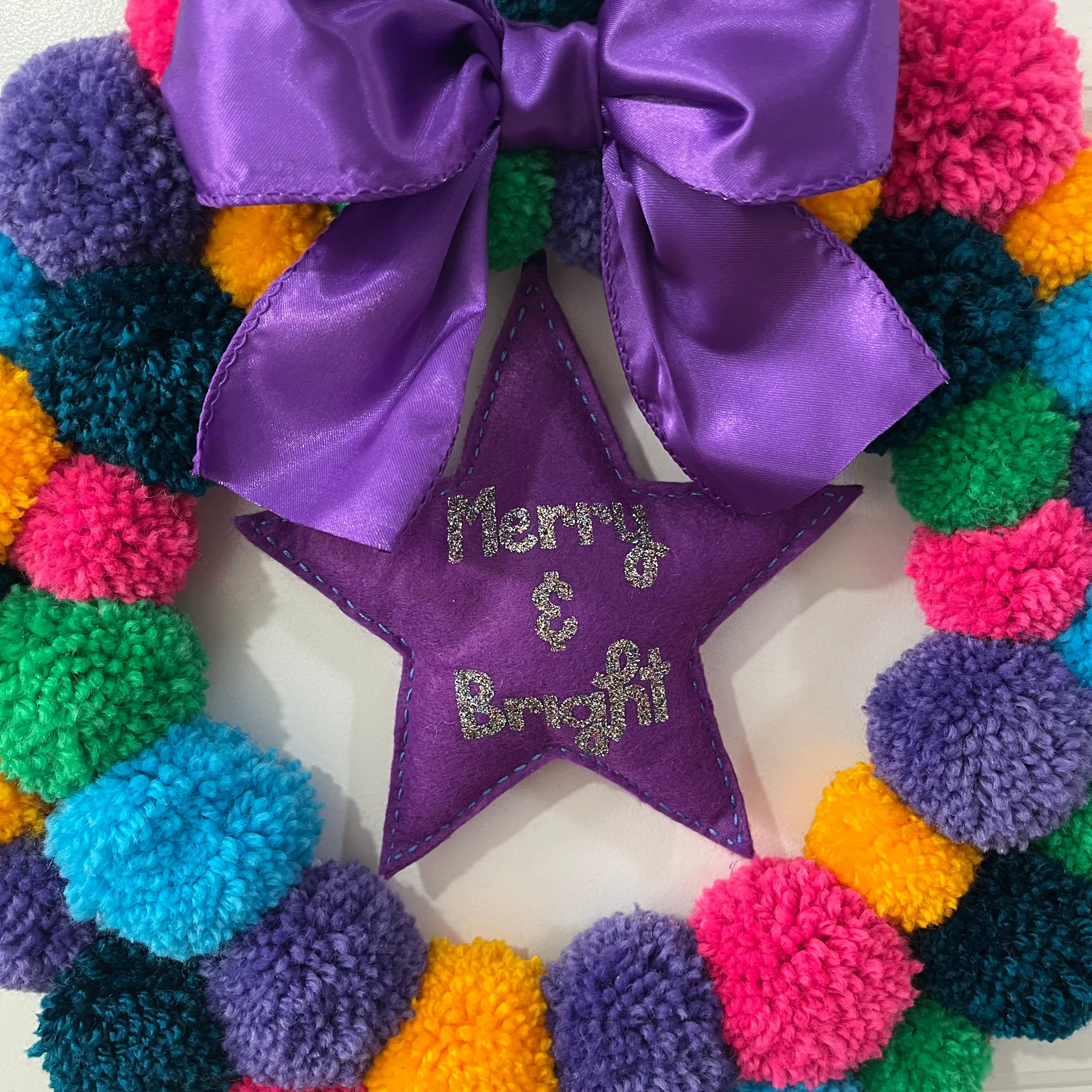 Close-up of a colorful pom-pom wreath with a purple bow and a star reading 'Merry & Bright'.