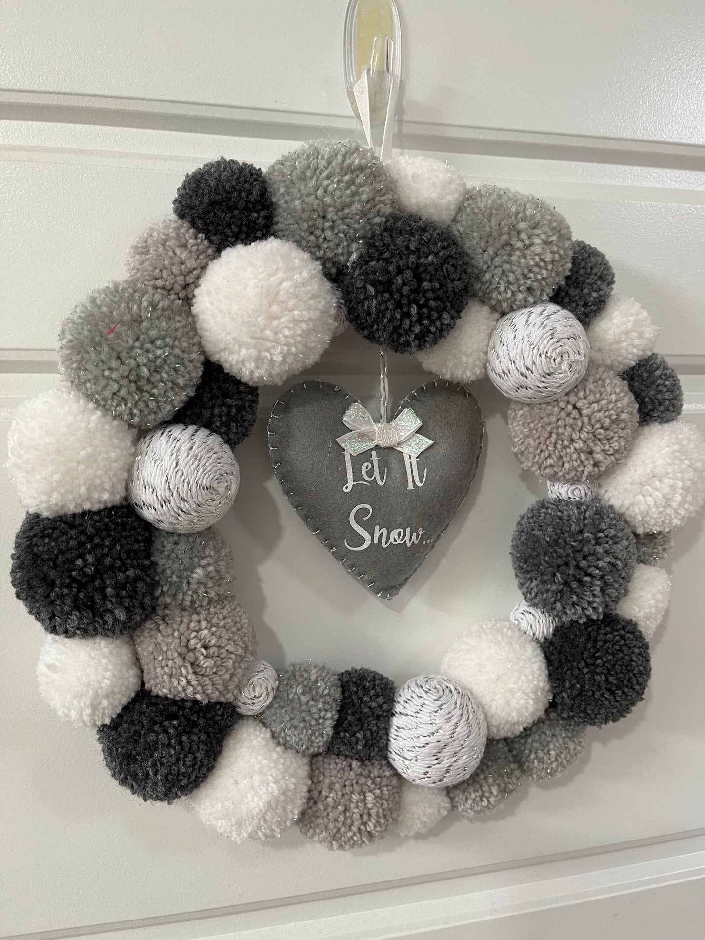 A festive wreath made of white, grey, and black pompoms with a heart-shaped 'Let It Snow' sign.