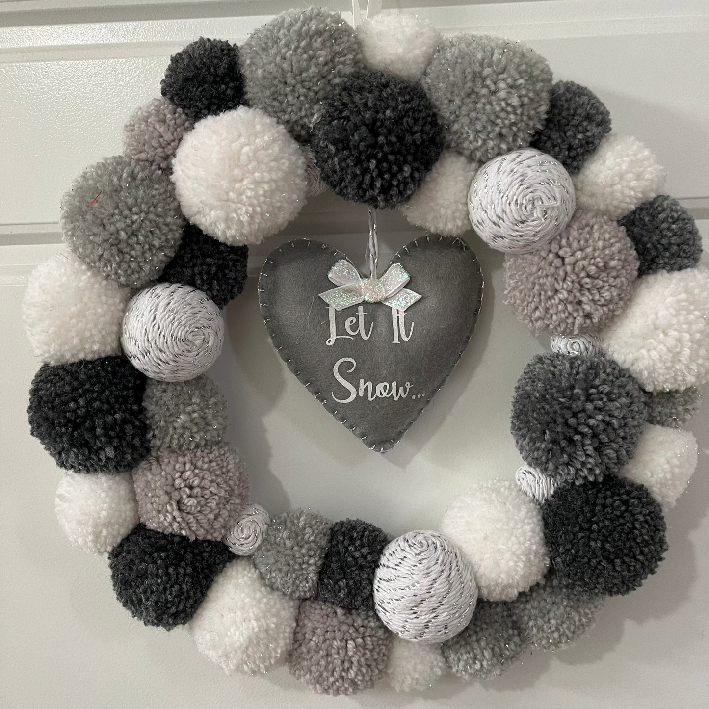A festive wreath made of white, grey, and black pompoms with a heart-shaped 'Let It Snow' sign.