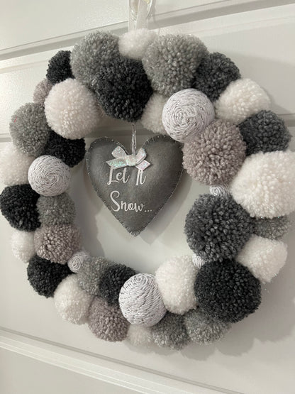 A festive wreath made of white, grey, and black pompoms with a heart-shaped 'Let It Snow' sign.