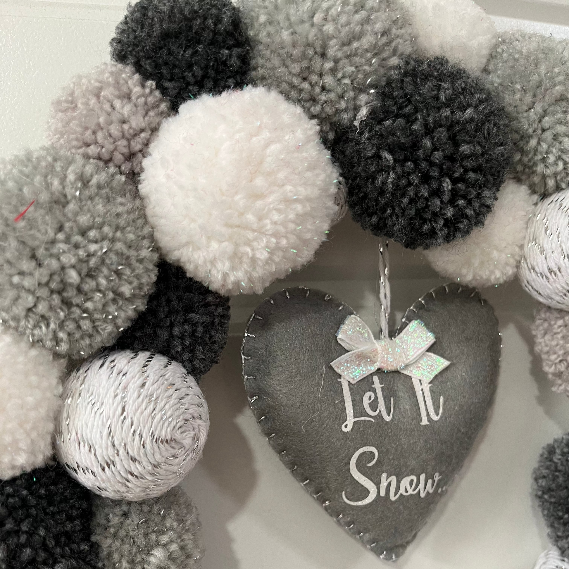 Close-up of a festive wreath with white, grey, black pompoms and a 'Let It Snow' heart.