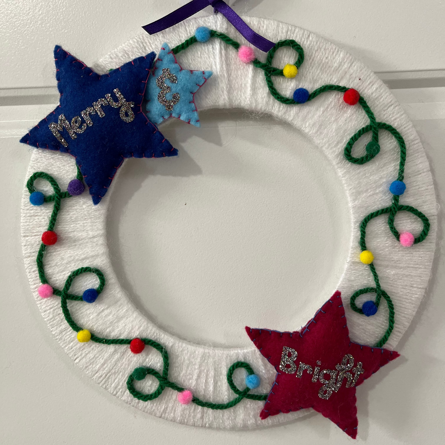 A white yarn-wrapped wreath adorned with colorful string lights and two felt stars saying "Merry" and "Bright".