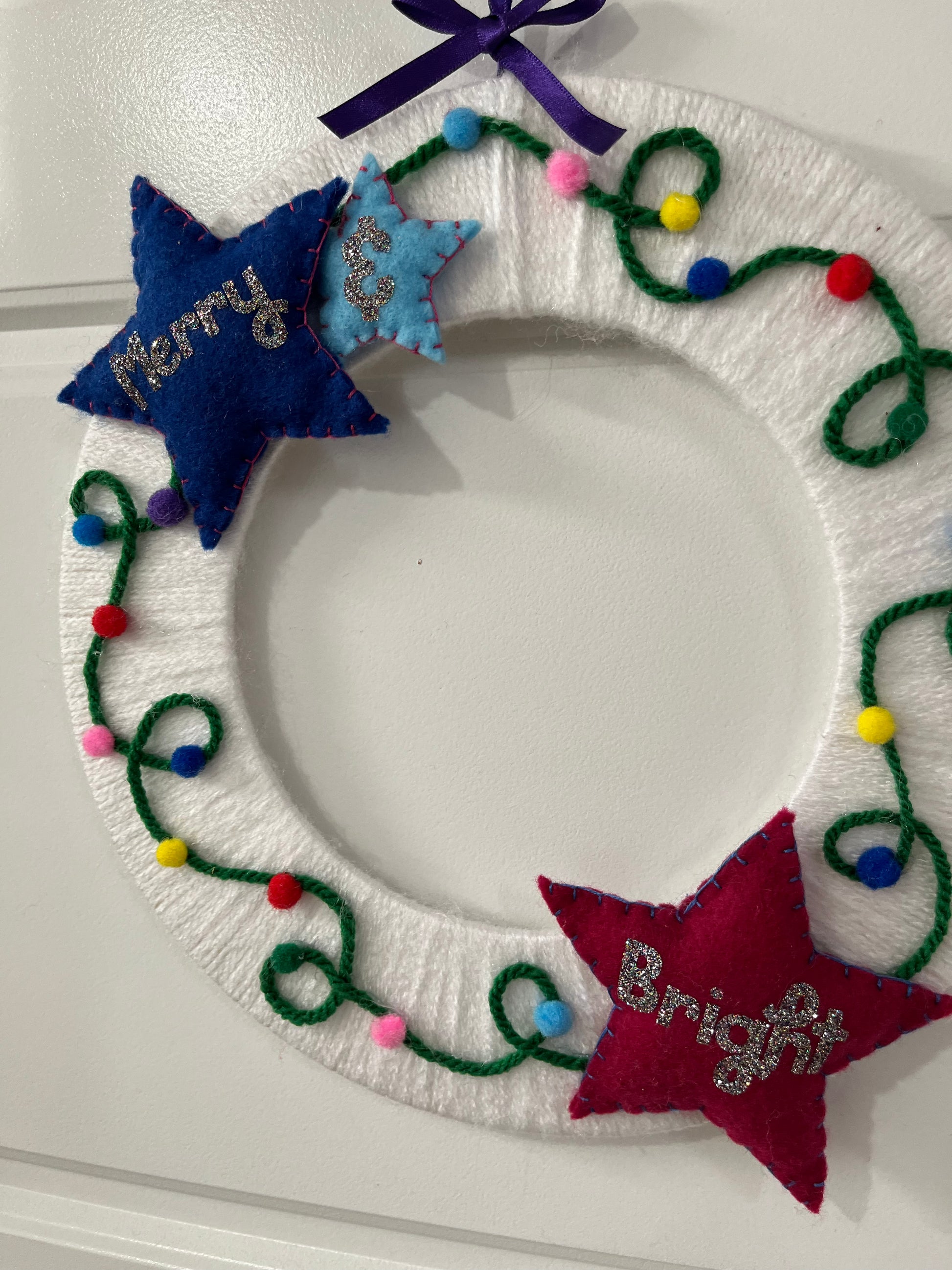 A festive white yarn wreath with colorful string lights and two felt stars, one blue, one red.
