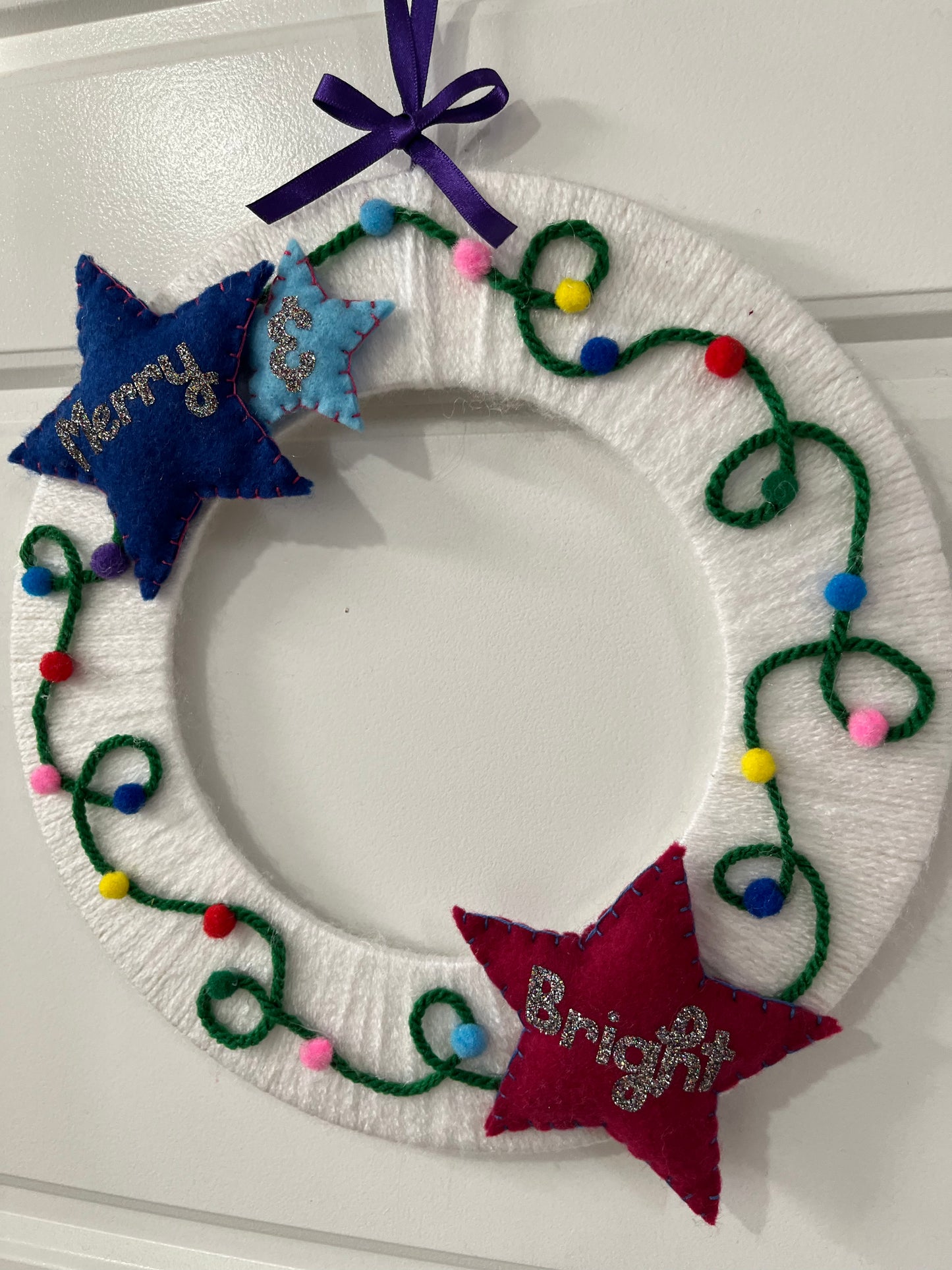 A white wreath decorated with string lights and two stars, one blue with "Merry" and one red with "Bright."