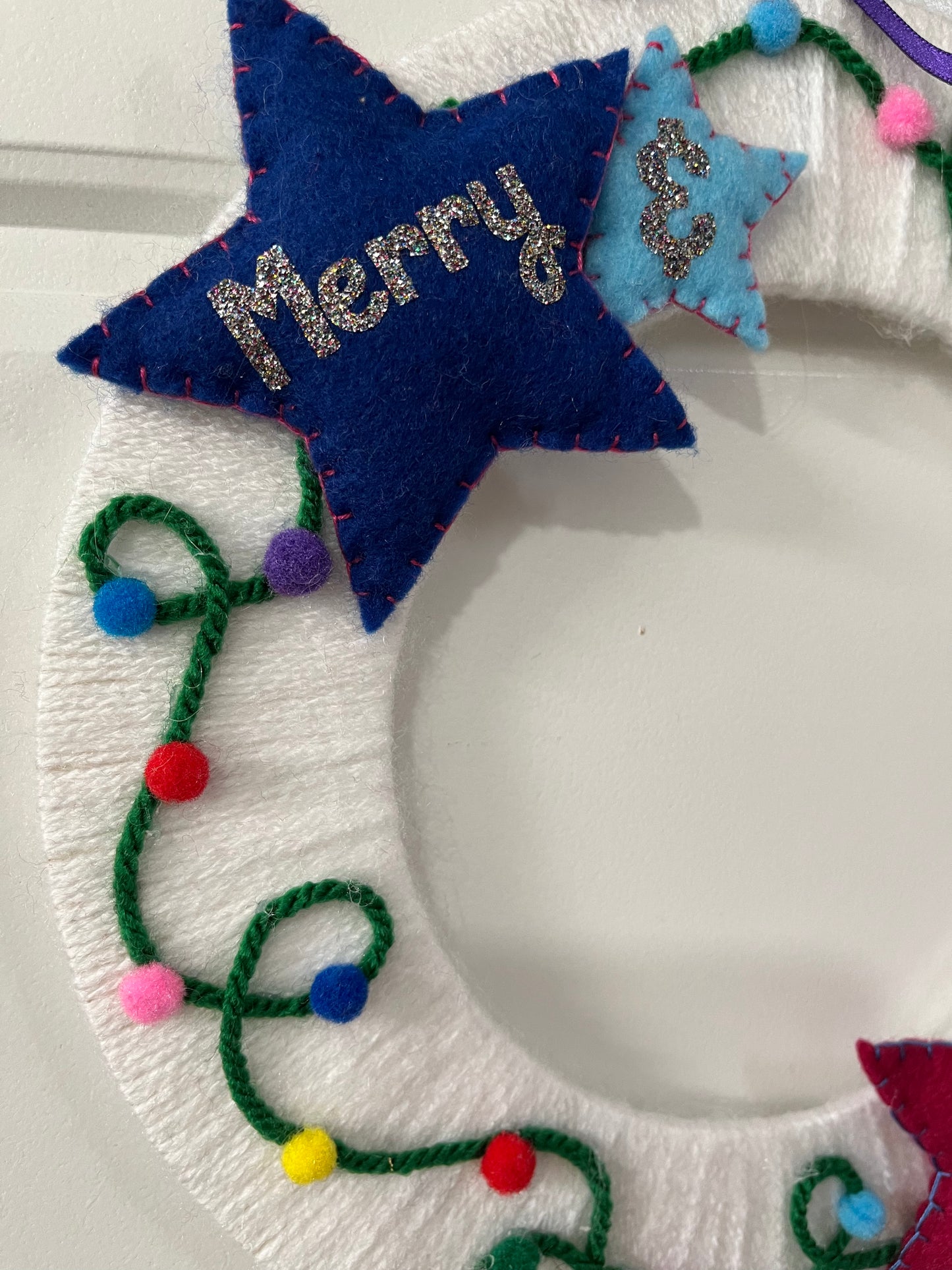 Close-up of a white wreath with a blue star saying "Merry" and colorful yarn lights.