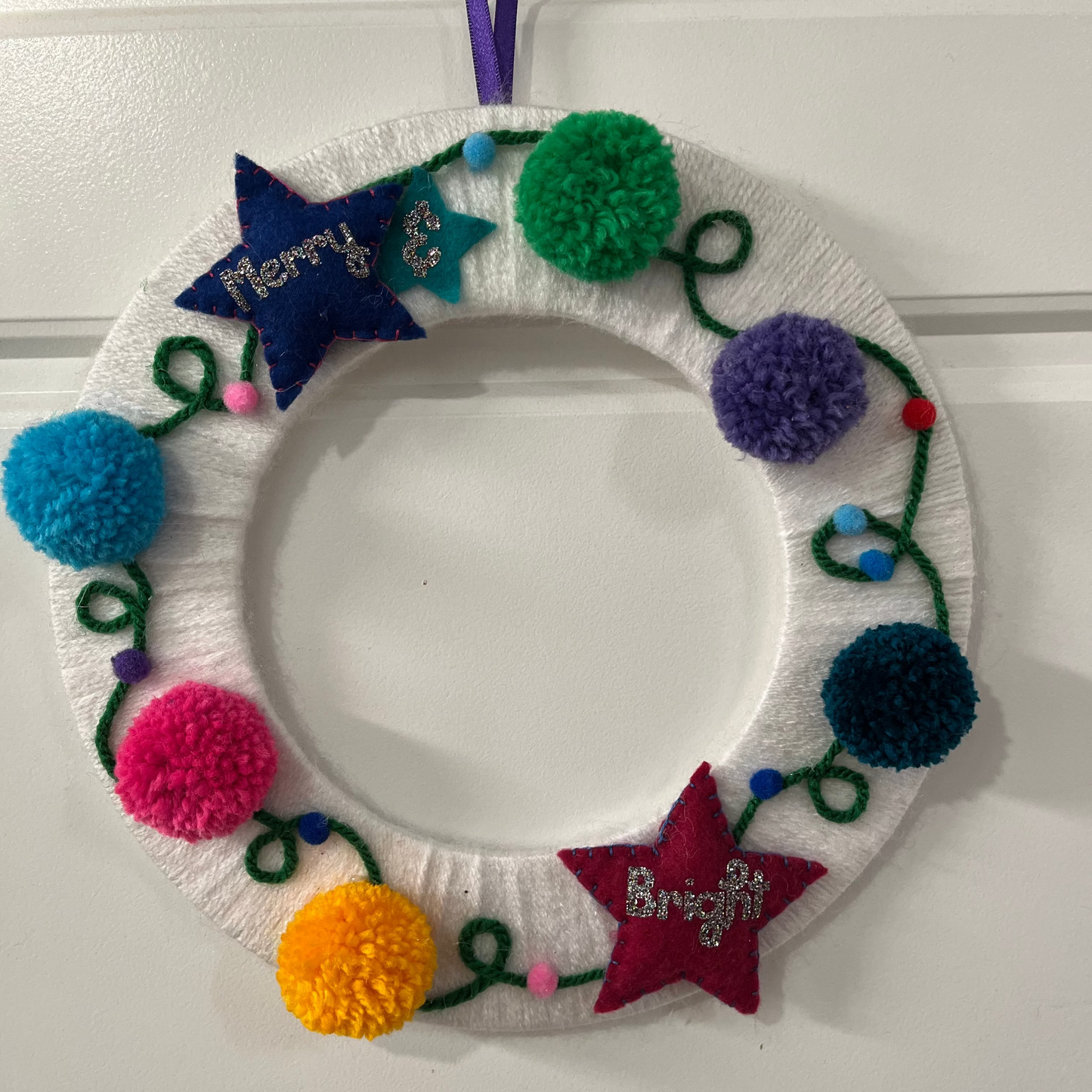 A white yarn-wrapped wreath adorned with colorful pom-poms, felt stars, and "Merry & Bright."