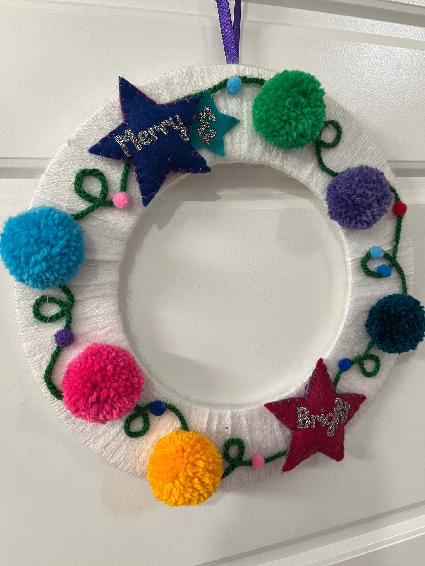 A white yarn-wrapped wreath adorned with colorful pom-poms, felt stars, and "Merry & Bright."