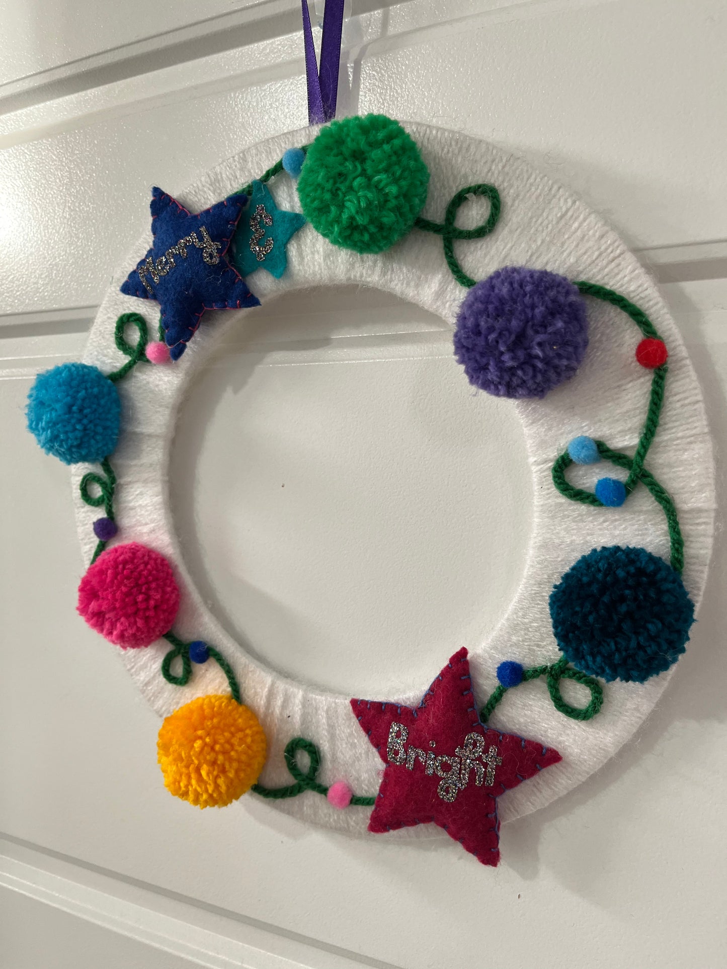A white yarn-wrapped wreath adorned with colorful pom-poms, felt stars, and "Merry & Bright."