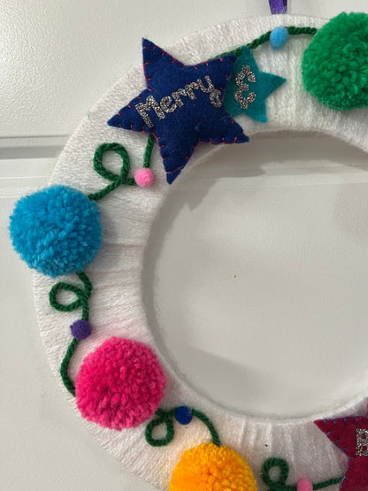 A white yarn-wrapped wreath adorned with colorful pom-poms, felt stars, and "Merry & Bright."