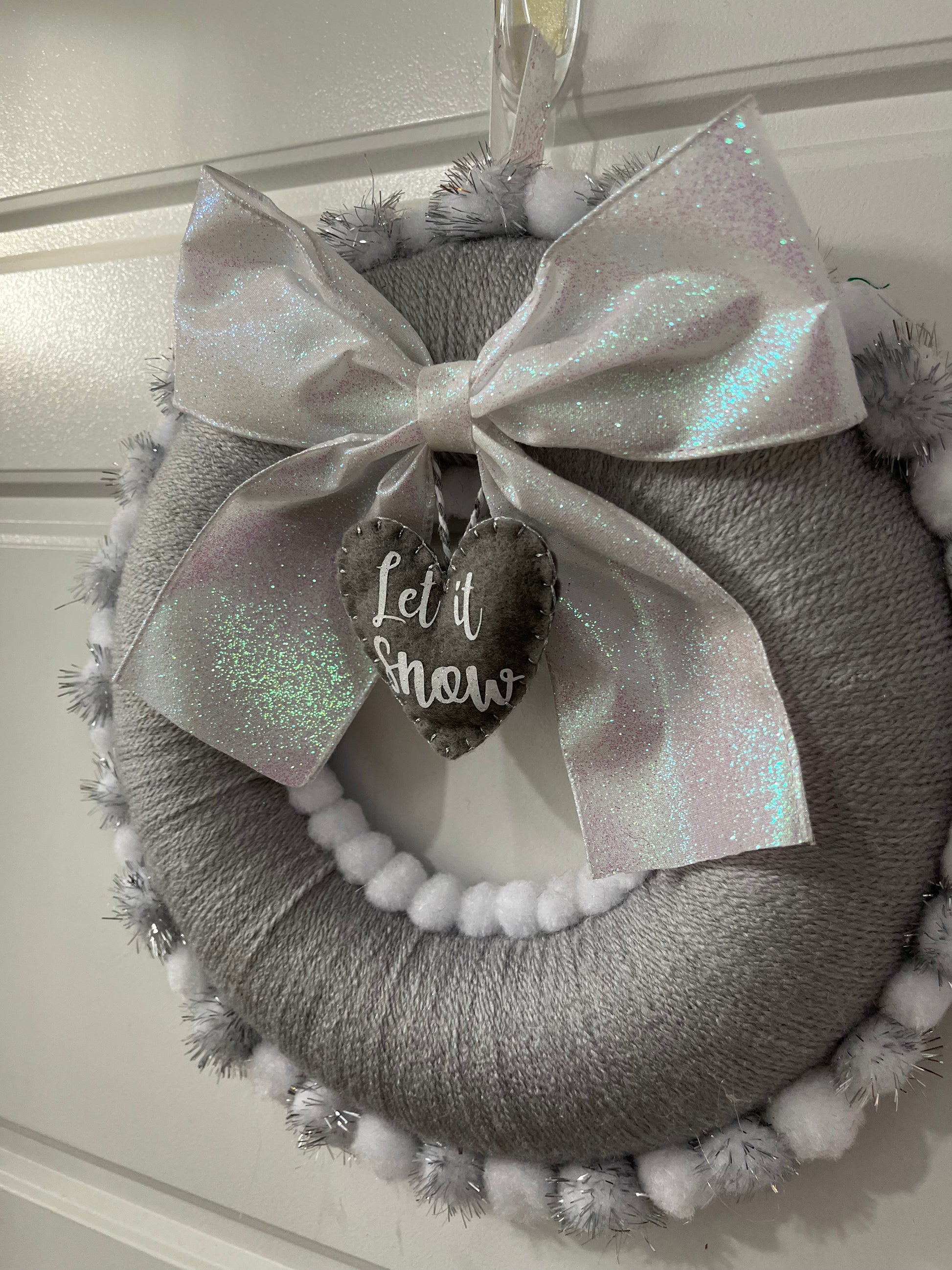 A festive gray wreath with a glittery white bow, a 'Let it Snow' tag, and fluffy trim.