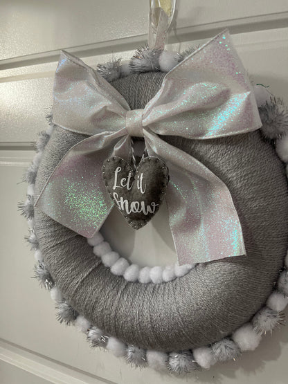 A festive gray wreath with a glittery white bow, a 'Let it Snow' tag, and fluffy trim.