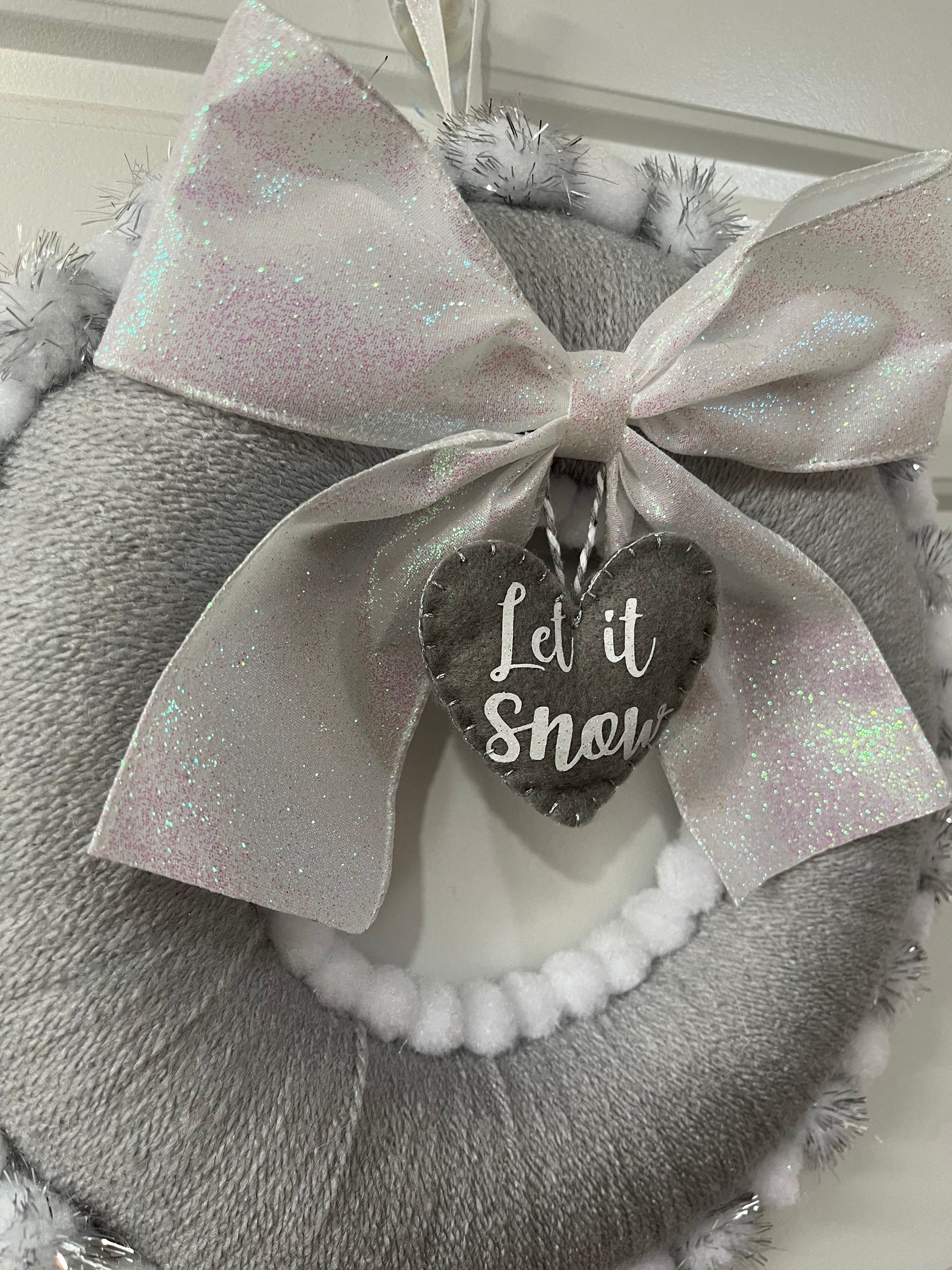 A close-up of a festive gray wreath with a glittery white bow and a heart-shaped 'Let it Snow' tag.