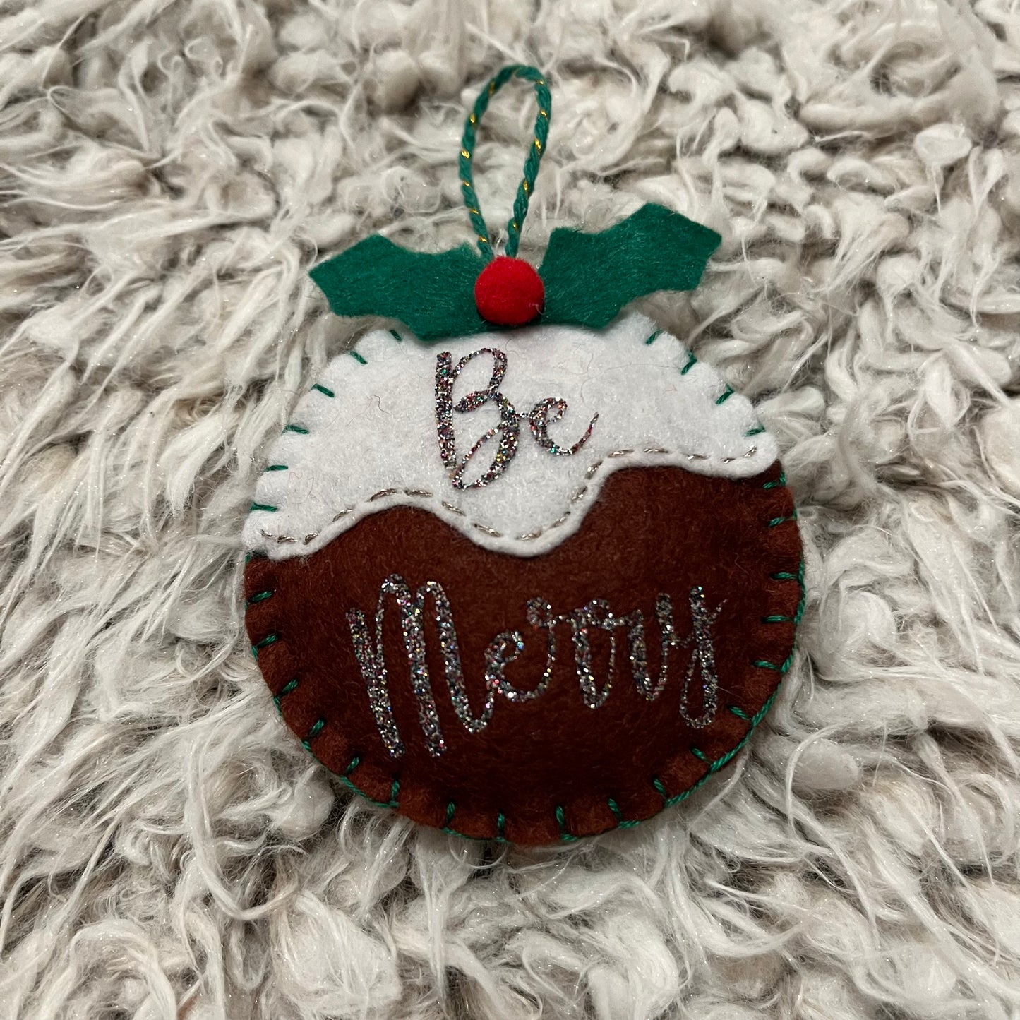 Handmade "Be Merry" Christmas pudding decoration on a white furry background.