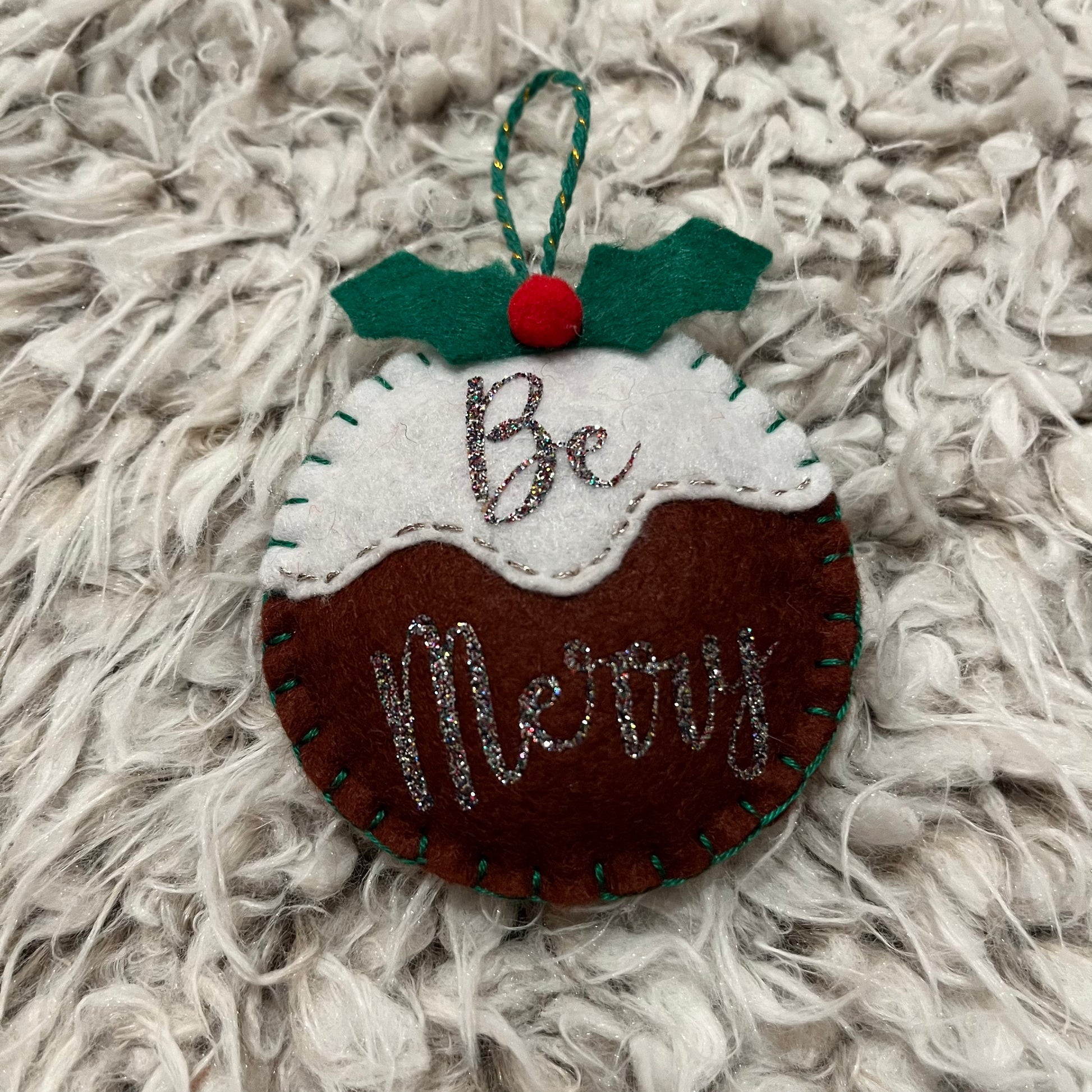Handmade "Be Merry" Christmas pudding decoration on a white furry background.