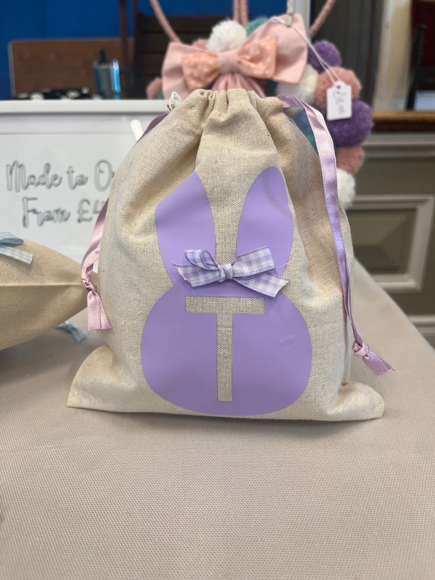 A beige drawstring bag with a purple bunny silhouette and the letter T, tied with a purple gingham bow.