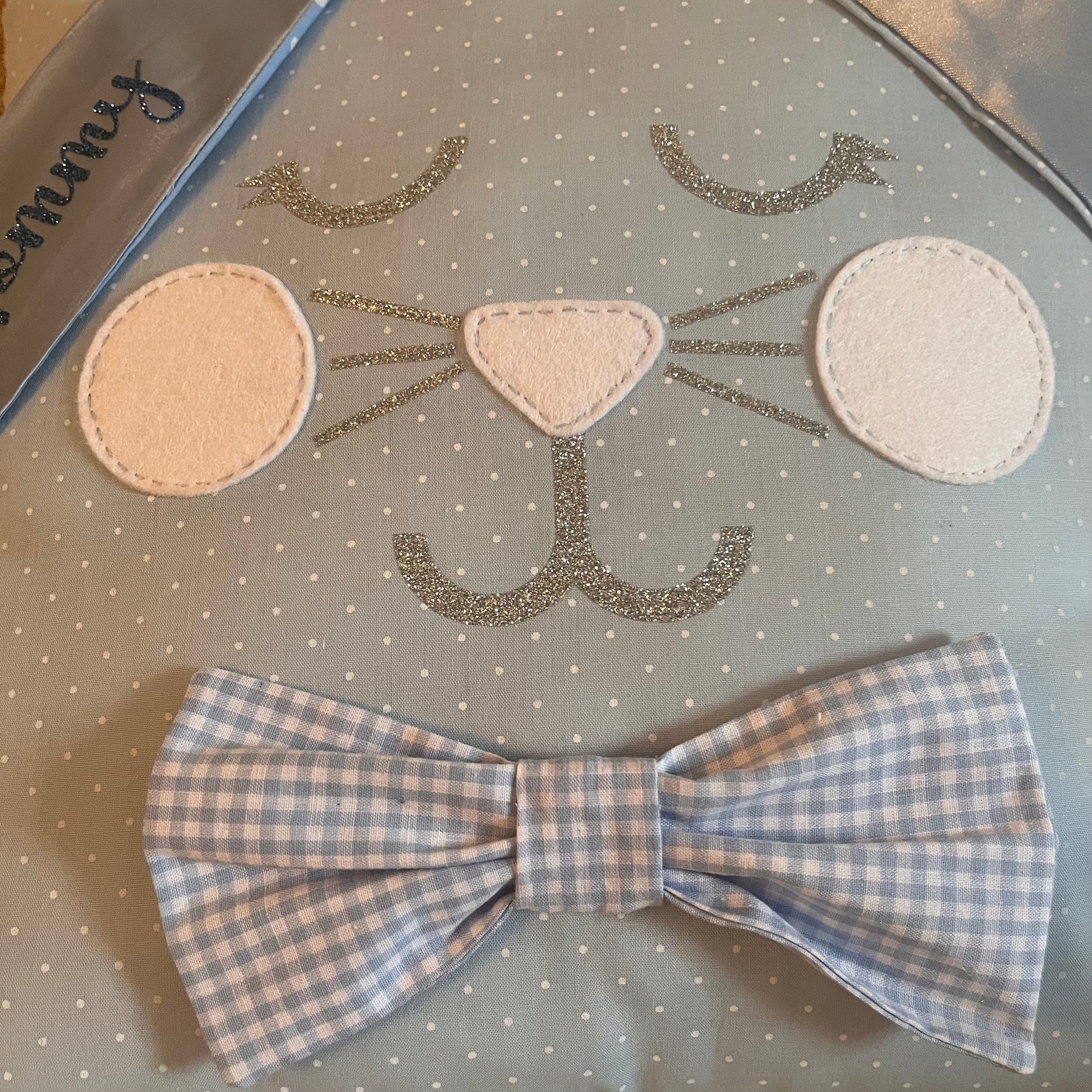 Close-up of a blue polka dot fabric with a glittery cat face and a blue gingham bow tie.