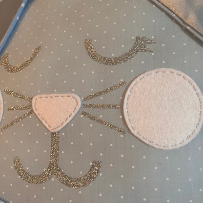 Close-up of a light blue polka dot fabric with a cat face design made of glitter and felt. The cat has a pink felt nose, glitter whiskers, and white felt eyes.