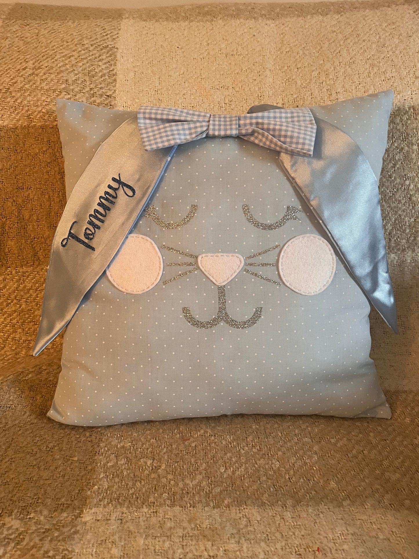 Light blue polka dot pillow with a bunny face, glittery whiskers, and a blue gingham bow. Tommy is embroidered on one ear.
