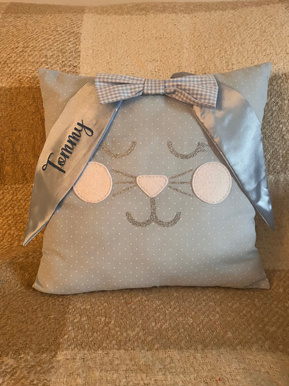 Light blue polka dot pillow with a bunny face, glittery whiskers, and a blue gingham bow. Tommy is embroidered on one ear.
