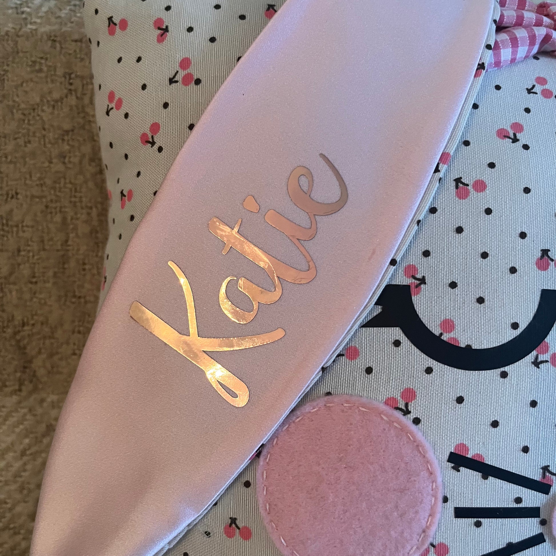 Close-up of a pink bunny ear headband with the name Katie in gold script, on a polka dot fabric.