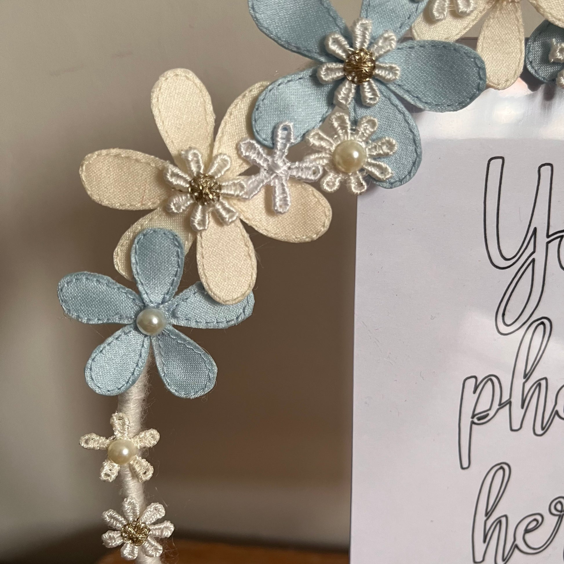 Close-up of a picture frame adorned with blue and cream fabric flowers, some with pearl or gold centers. 
