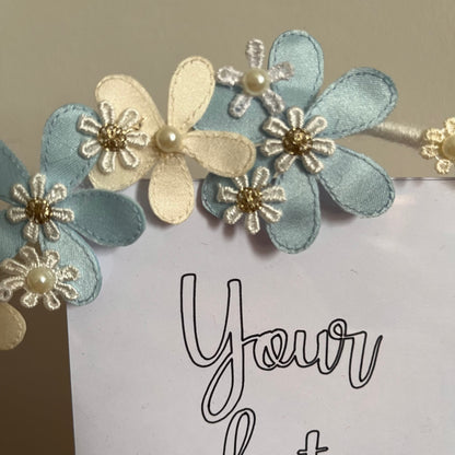 Close-up of a picture frame adorned with light blue and cream fabric flowers, some with pearl centers.