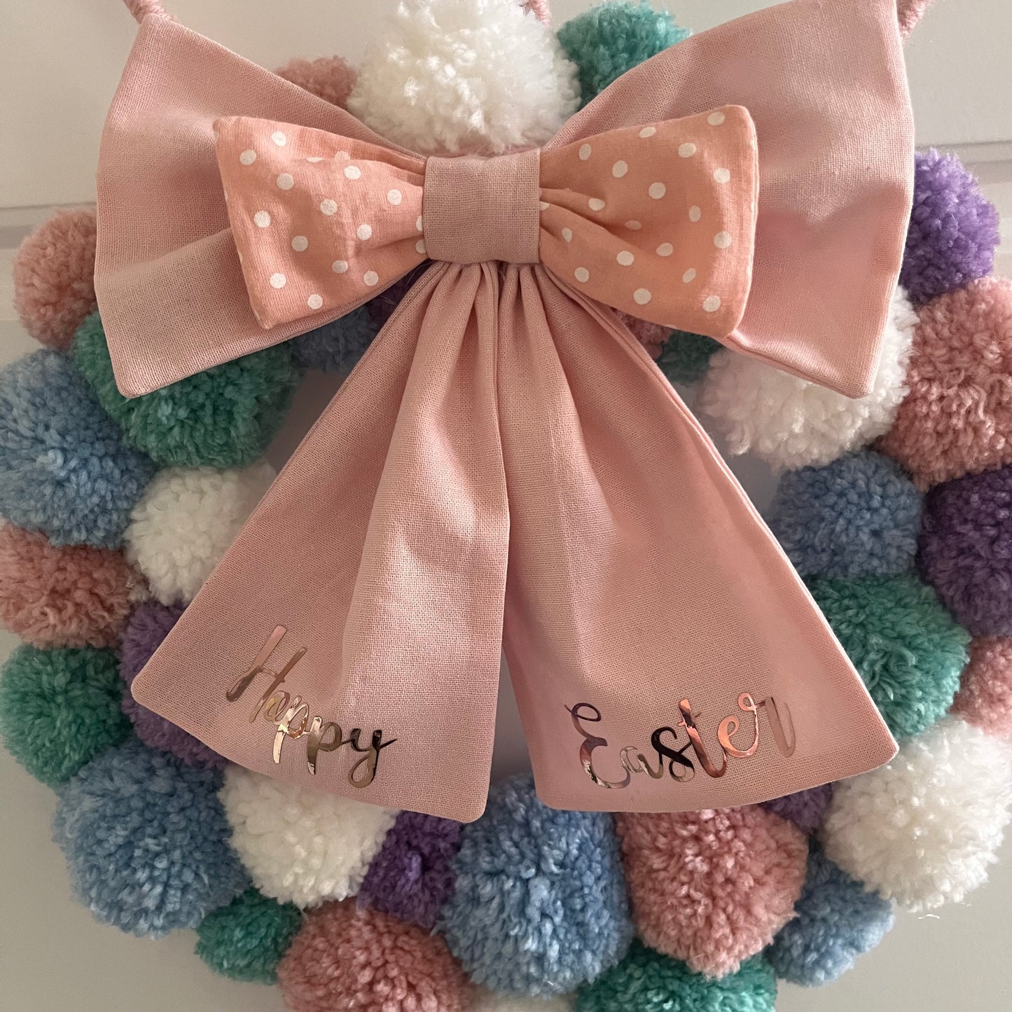 A pastel pom-pom wreath with a pink bow that says Happy Easter.