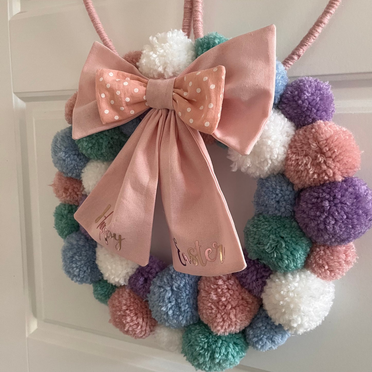 A pastel pom-pom wreath with a pink polka-dot bow that reads Happy Easter.
