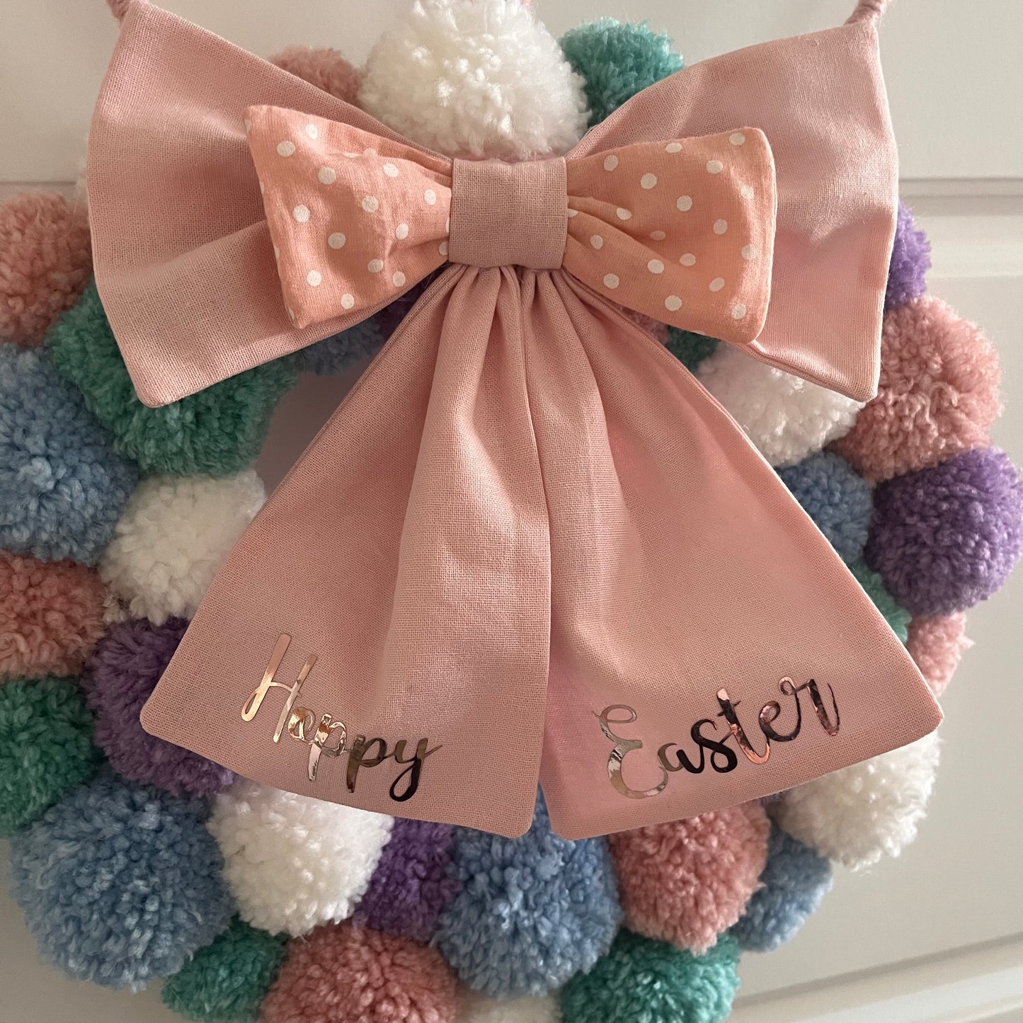 Pastel pom-pom wreath with a pink bow that says Happy Easter in gold lettering.