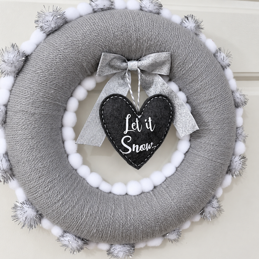 Gray yarn wreath with white pom-poms and silver tinsel. A silver bow and black heart with Let it Snow.