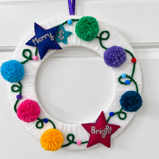 A white yarn-wrapped wreath adorned with colorful pom-poms, felt stars, and "Merry & Bright."