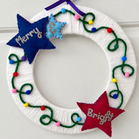 A white yarn-wrapped wreath adorned with colorful string lights and two felt stars saying "Merry" and "Bright".