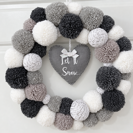 A festive wreath made of white, grey, and black pompoms with a heart-shaped 'Let It Snow' sign.