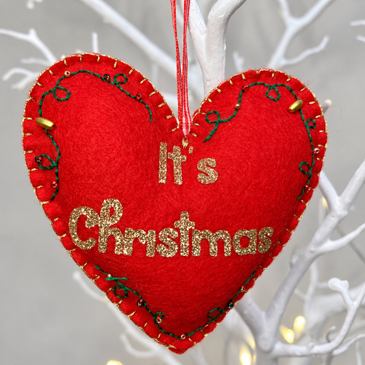 Red felt heart ornament with gold glitter text It's Christmas, green embroidery, and gold beads. Hanging on a white branch.