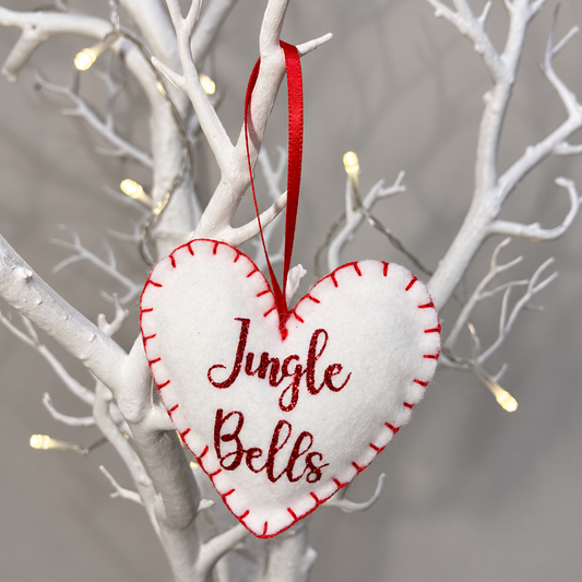A white felt heart ornament with Jingle Bells in red glitter, hanging from a branch.