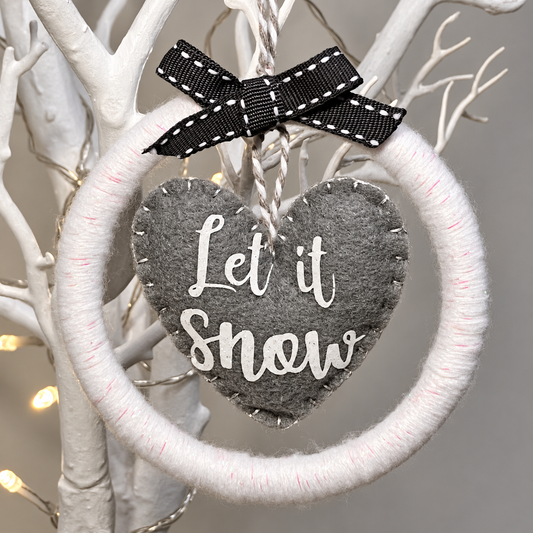 A white yarn wreath with a gray felt heart saying Let it Snow.