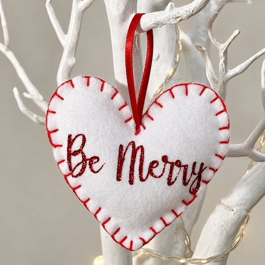 White felt heart ornament with red stitching and "Be Merry" in glitter script.