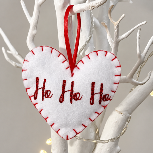 White felt heart ornament with red stitching and "Ho Ho Ho" in glitter, hanging on a white branch.