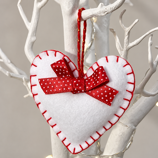 White felt heart ornament with red stitching and a red polka dot bow, hanging on a white branch.