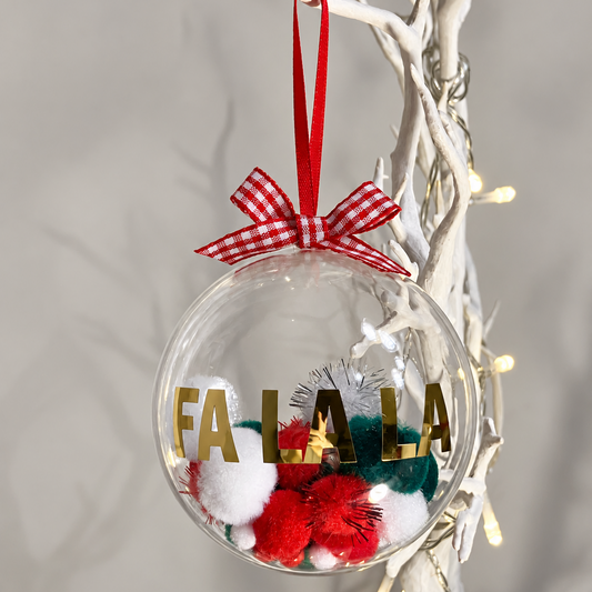 Clear Christmas bauble with "FA LA LA" in gold, filled with red, white, and green pom-poms.