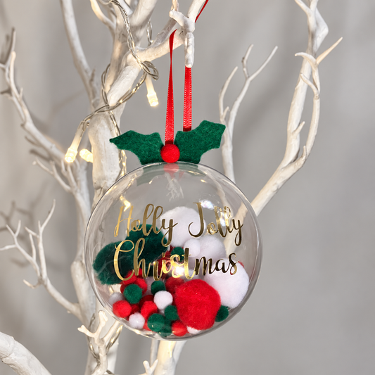 Clear Christmas bauble with green and red pom-poms and gold lettering.