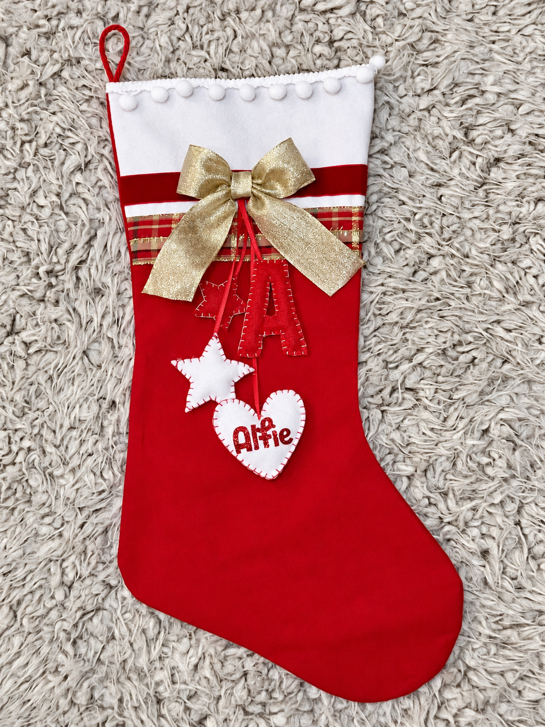 Red Christmas stocking with a gold bow, a white star, and a heart with "Alfie" written on it.