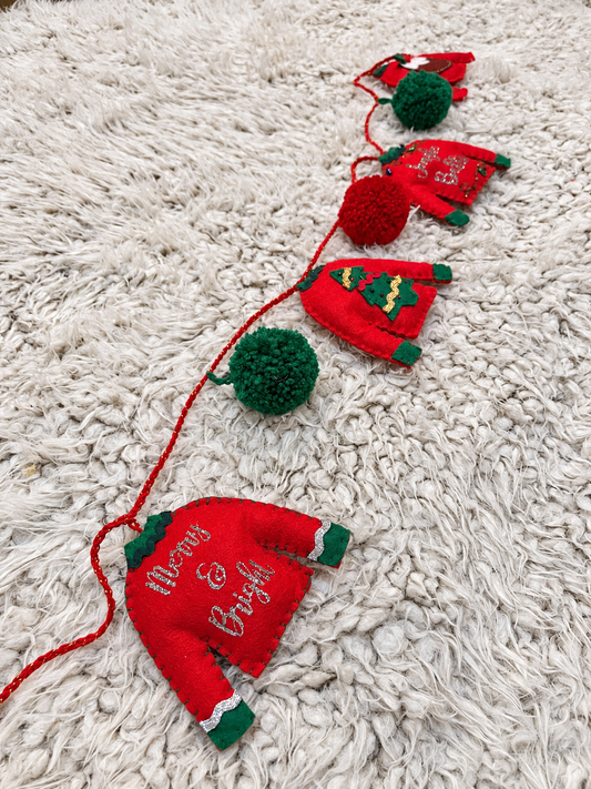 Red Christmas sweater garland with green pom-poms on a white fluffy blanket.
