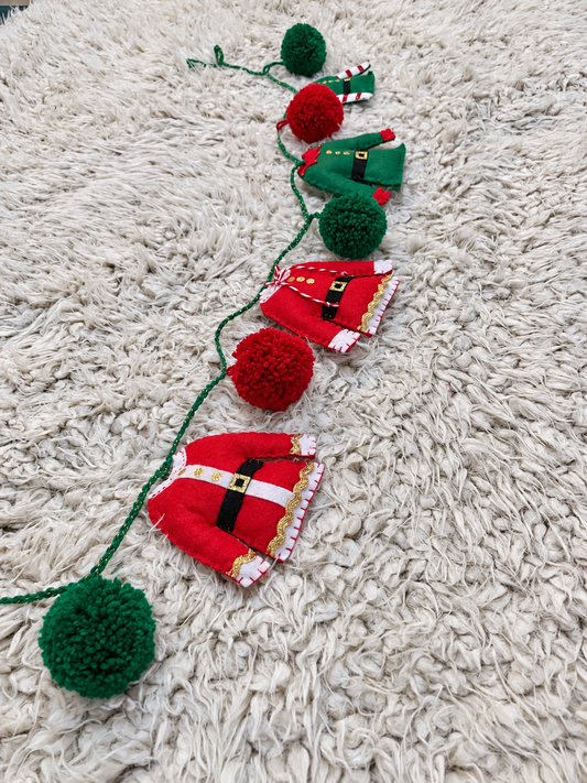 Christmas garland with red Santa suits, green elf outfits, and red and green pom-poms on a white rug.