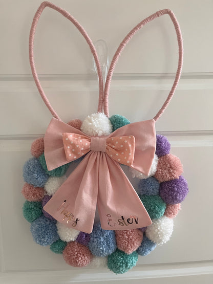 Easter bunny wreath with pastel pom-poms and a pink bow that says Happy Easter.