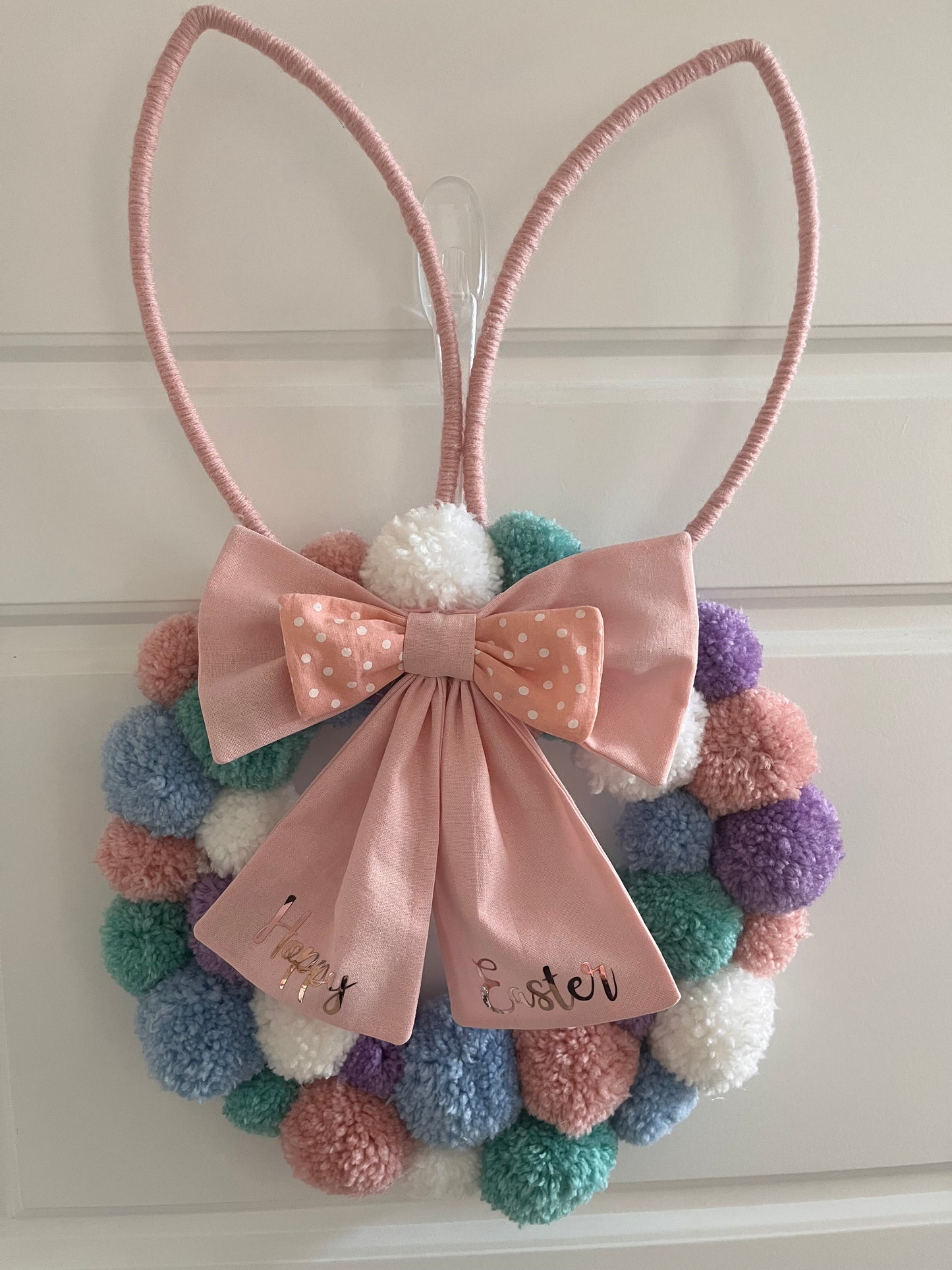 Easter bunny wreath with pastel pom-poms and a pink bow that says Happy Easter.
